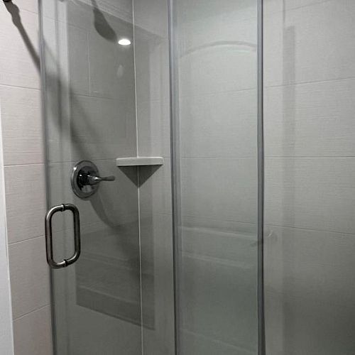 The image shows a modern shower with a sliding glass door and a metal handle; there is a showerhead and a small shelf visible inside.