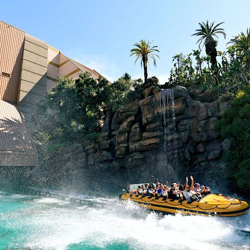 A group of people is on a yellow water ride with splashes in an amusement park, surrounded by lush greenery and a waterfall, under a bright sky.