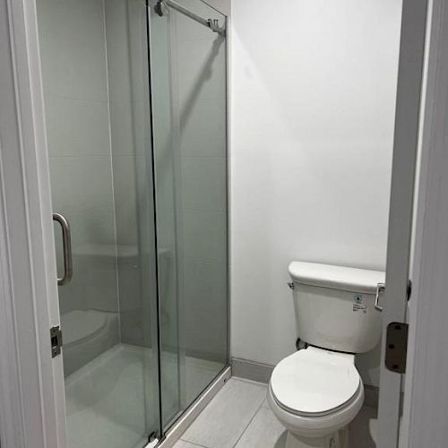 A small bathroom with a glass-enclosed shower on the left and a white toilet on the right, featuring white tiled flooring and walls.