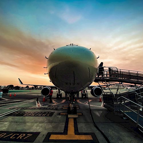 The image shows the front view of an airplane on the tarmac at sunset, with boarding stairs leading up to it and airport markings on the ground.