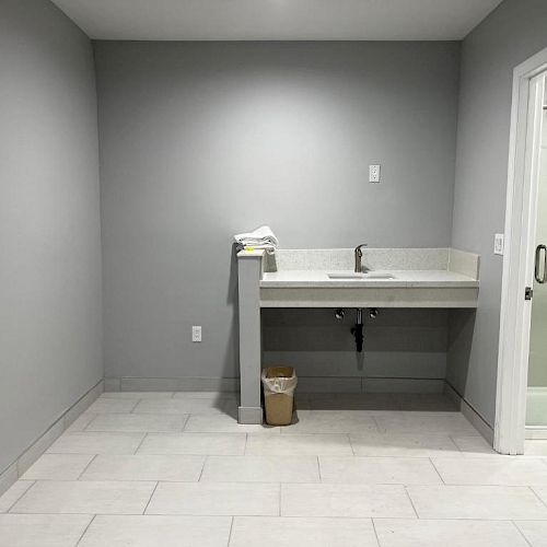 A minimalist room with gray walls, a sink with countertop, a trash bin underneath, and a partially visible glass door to the right.