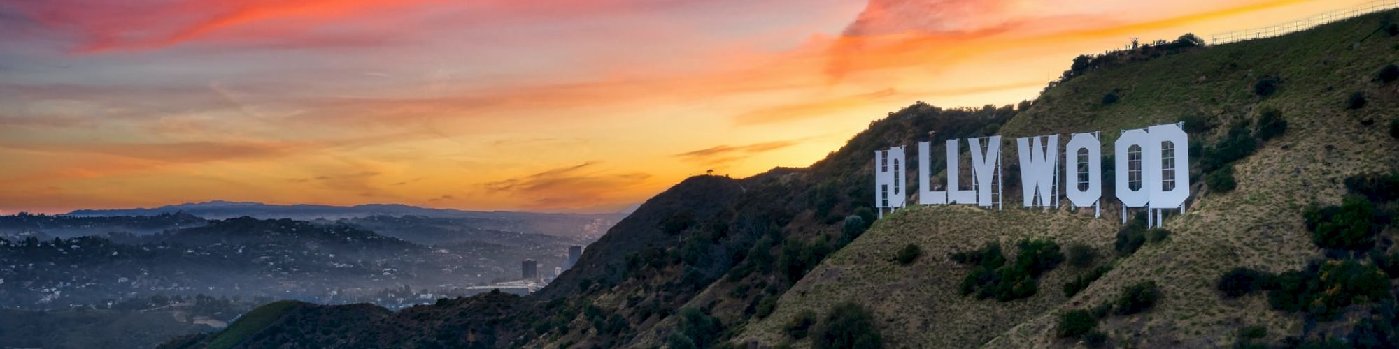 The image shows the iconic Hollywood sign on a hillside at sunset, with a colorful sky and the cityscape of Los Angeles in the background.