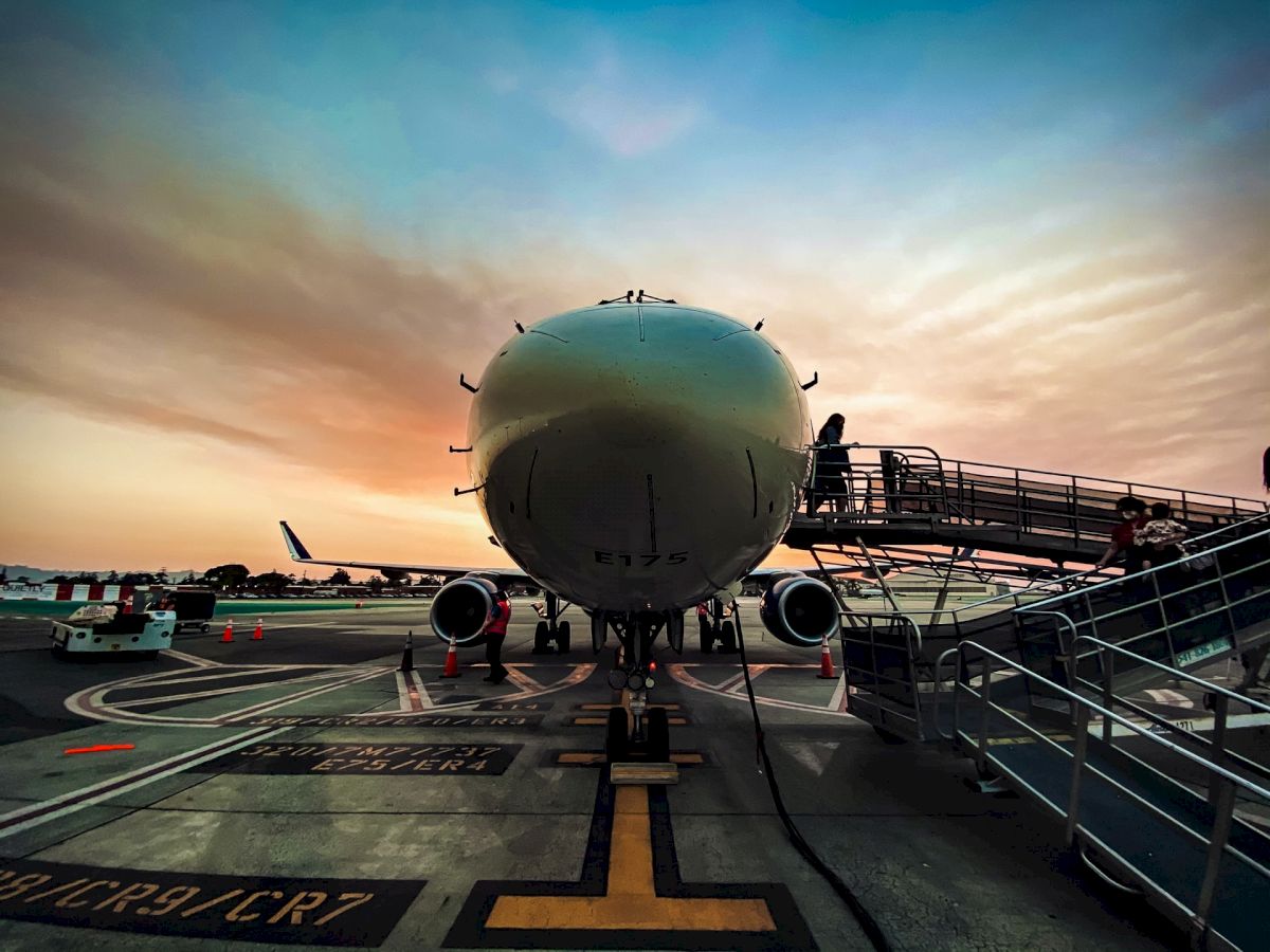 An airplane is parked on the tarmac under a colorful sky, with a boarding ramp extending to its entrance and ground support equipment in view.