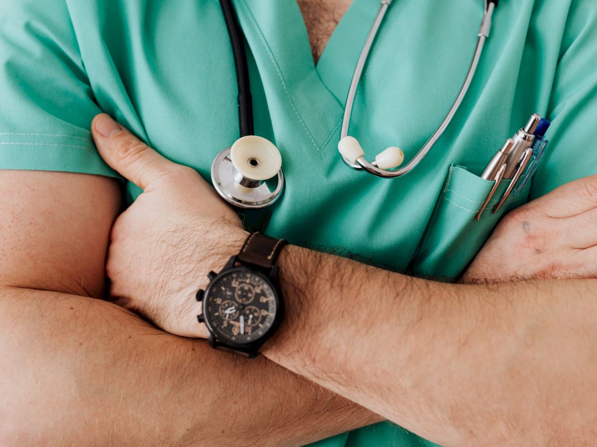 A person in green scrubs with a stethoscope around their neck, arms crossed, and pens in the pocket, likely a medical professional.