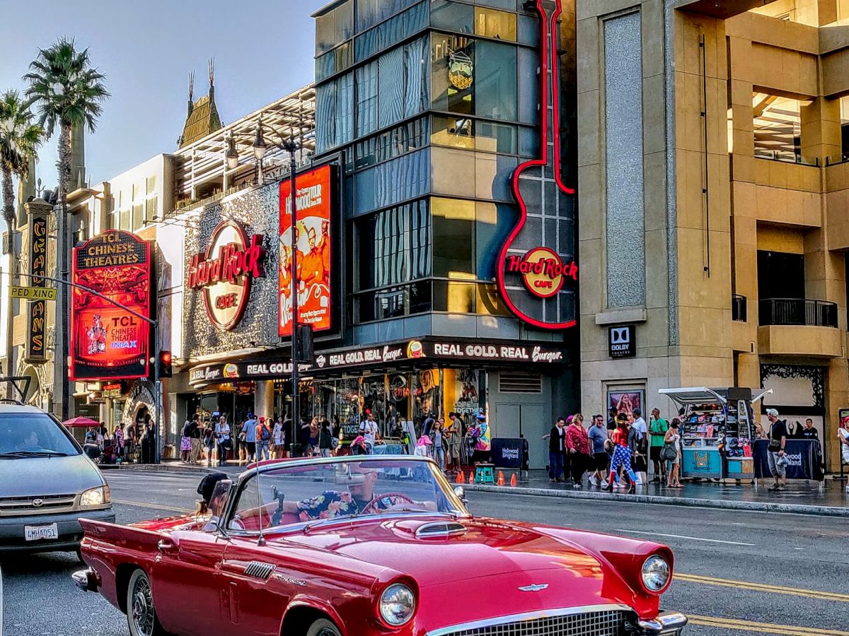 A red vintage car drives down a busy street with neon signs, shops, and a theater in the background. Palm trees line the sidewalk.