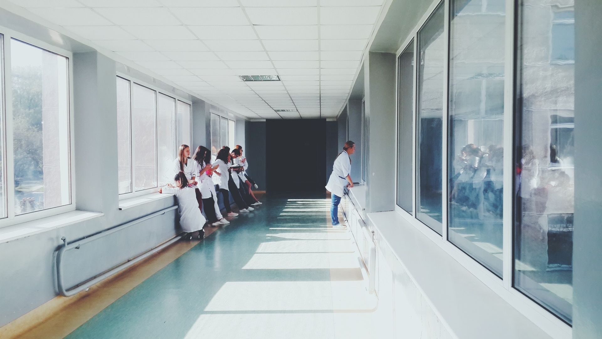 A group of people in white coats stand together in a brightly lit hallway with large windows, while one person walks alone on the other side.