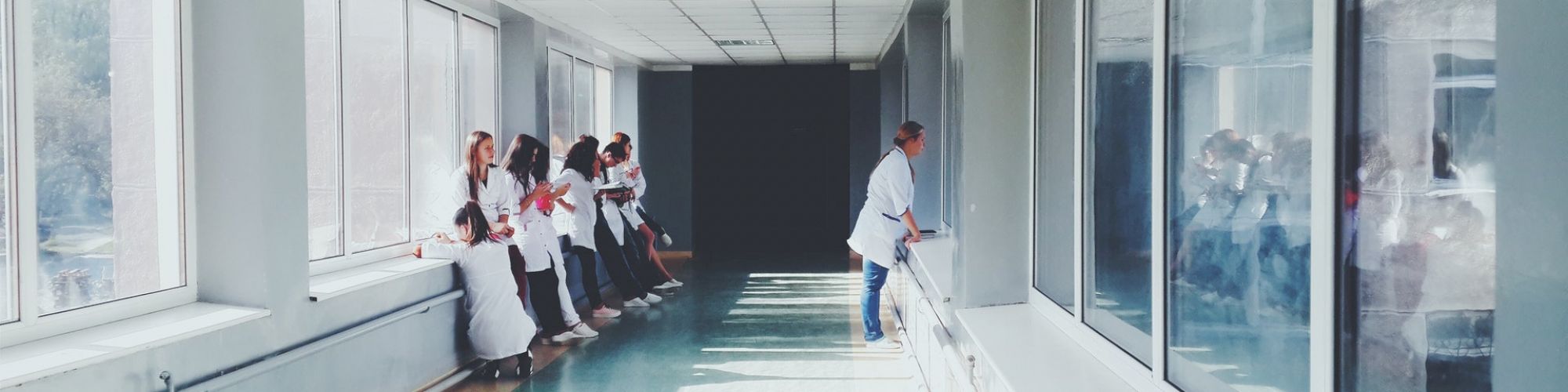 A group of people in white coats stand together in a brightly lit hallway with large windows, while one person walks alone on the other side.