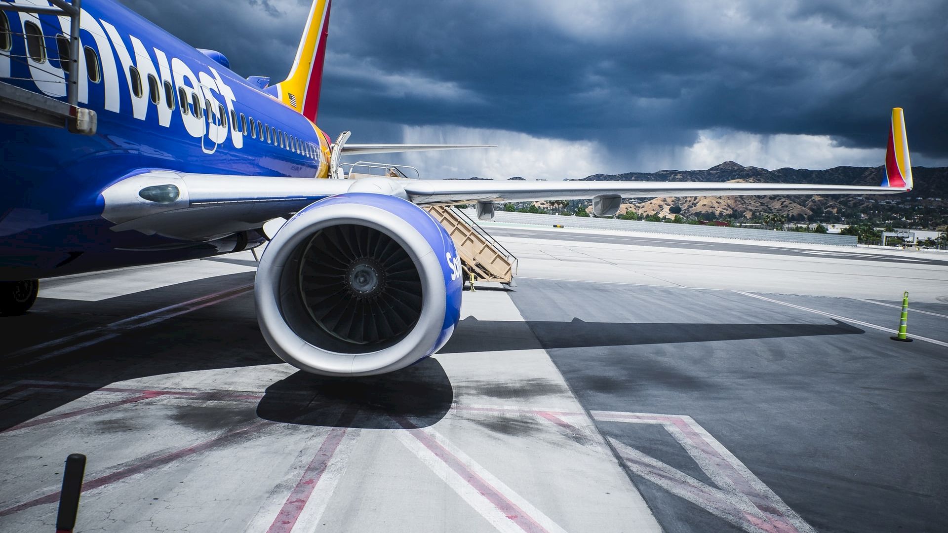 A close-up of a commercial airplane on a runway with a partly cloudy sky and mountains in the background, showcasing its engine and wing.