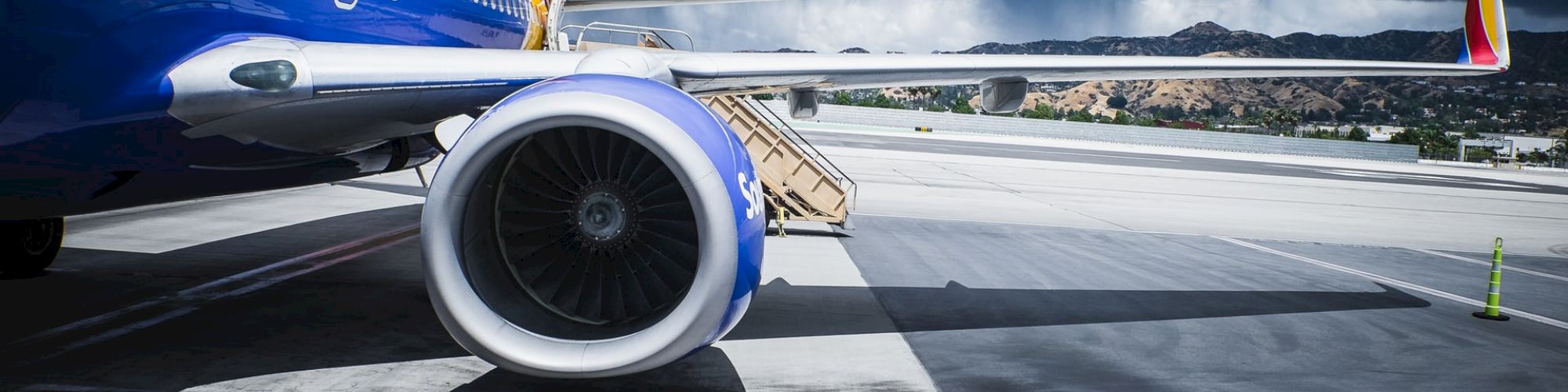 A close-up of a commercial airplane on a runway with a partly cloudy sky and mountains in the background, showcasing its engine and wing.