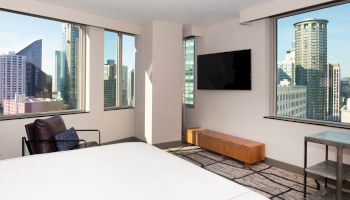 A modern hotel room with large windows showing a cityscape, a wall-mounted TV, chair, and minimalist furniture.