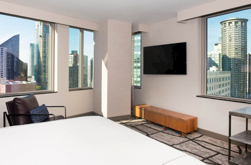 A modern hotel room with large windows showing a cityscape, a wall-mounted TV, chair, and minimalist furniture.