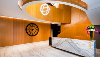 A hotel lobby features a modern reception desk, a large wall clock, spiral staircase, and floral arrangements in a well-lit space.