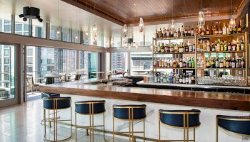 The image shows a modern bar with a long counter, high stools, a fully stocked liquor shelf, and large windows with a city view in the background.