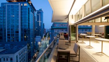 A modern urban balcony overlooks a city street with tall buildings, featuring outdoor seating and tables, and illuminated by city lights.
