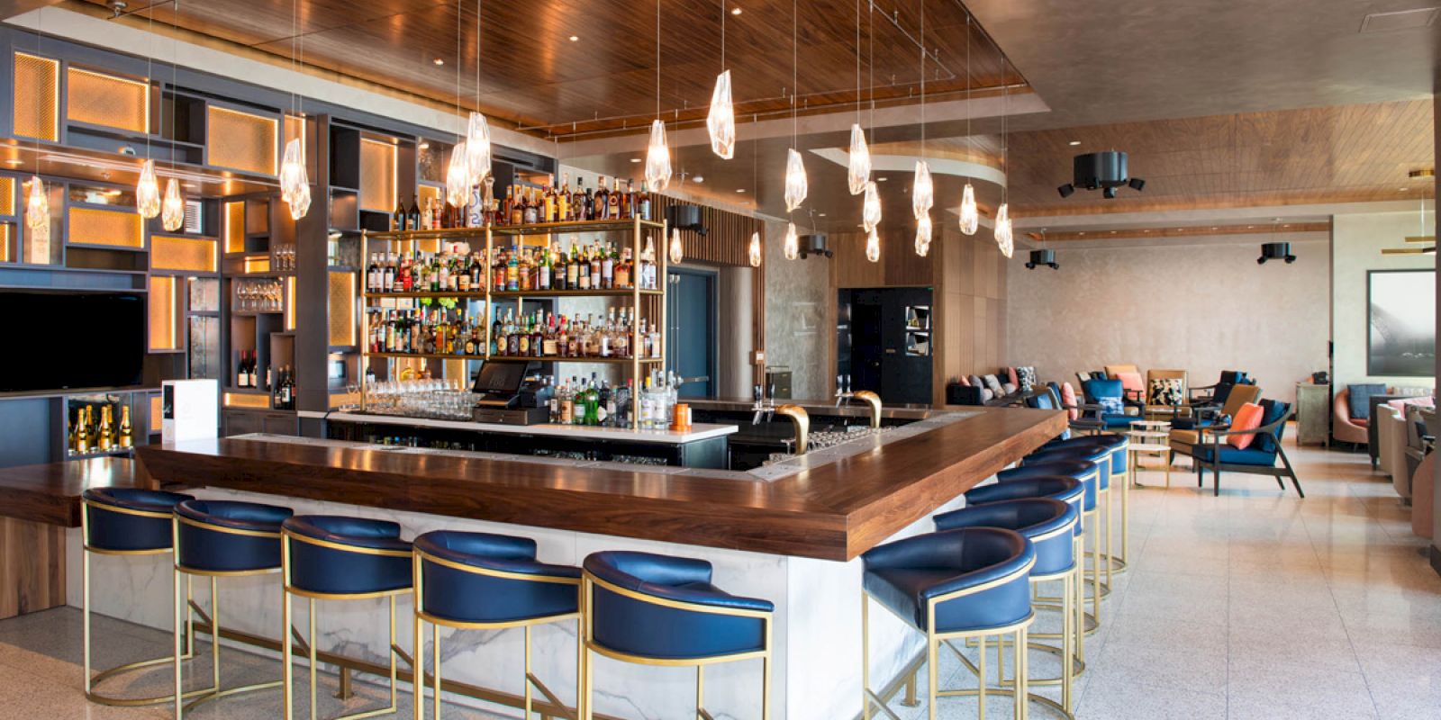 A modern bar with a wooden counter, high chairs, hanging lights, and a well-stocked shelves of bottles. The seating area is in the background.