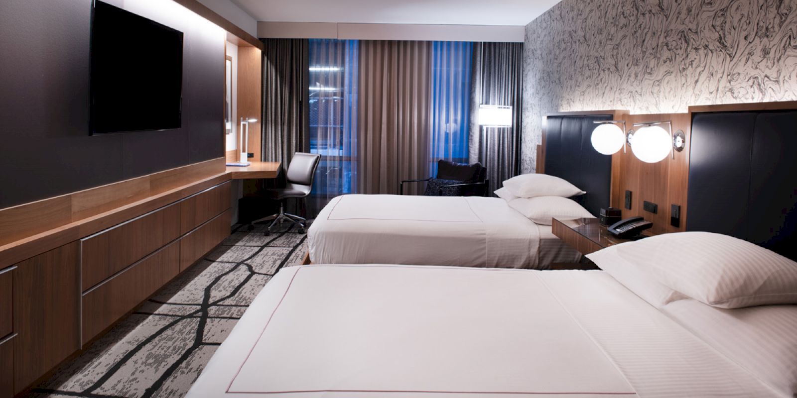 A modern hotel room with two beds, wall-mounted TV, desk, and chair, featuring contemporary decor and large windows with curtains.