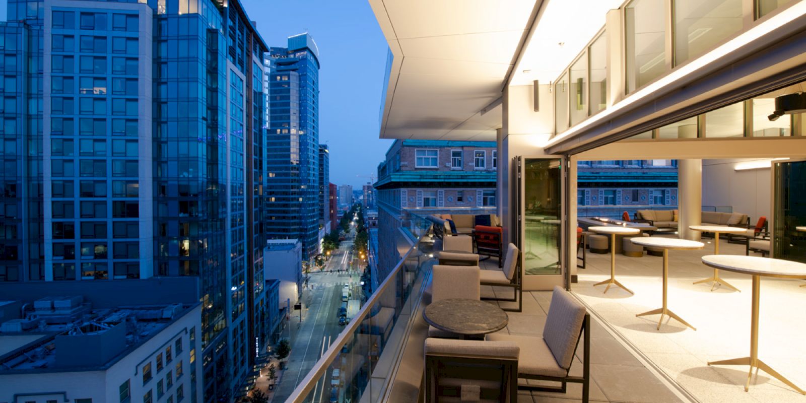 The image shows a modern balcony at dusk with chairs and tables, overlooking a city skyline of tall buildings and a busy street below.