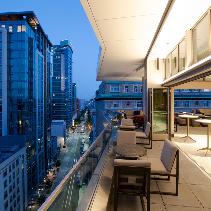 A modern cityscape view with tall buildings is seen from a rooftop terrace featuring tables, chairs, and soft ambient lighting in the evening.