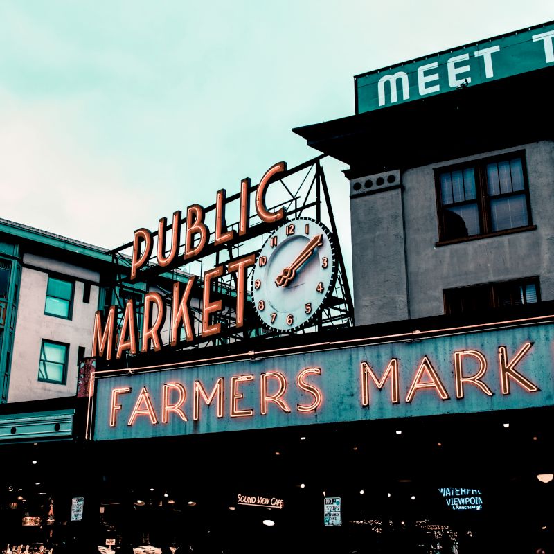 The image depicts the famous Pike Place Market in Seattle, featuring its iconic "Public Market" sign and a clock, with "Farmers Market" in neon lights.