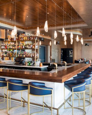The image shows a modern bar with a wooden counter, blue chairs, an assortment of bottles, pendant lights, and a cozy seating area in the background.