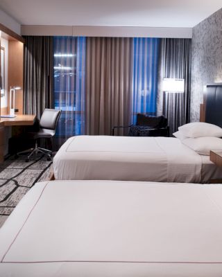 The image shows a modern hotel room with two neatly made beds, a wall-mounted TV, a desk, and floor-to-ceiling windows with drapes.