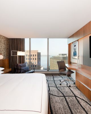 A modern hotel room with a king-sized bed, large flat-screen TV, desk, chair, and a view of buildings and water through large windows.