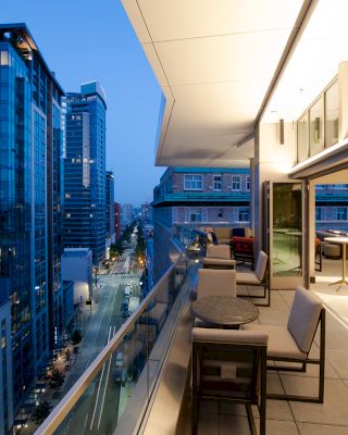A modern balcony overlooking a cityscape at dusk, featuring outdoor seating and tables, with buildings and illuminated street below it.