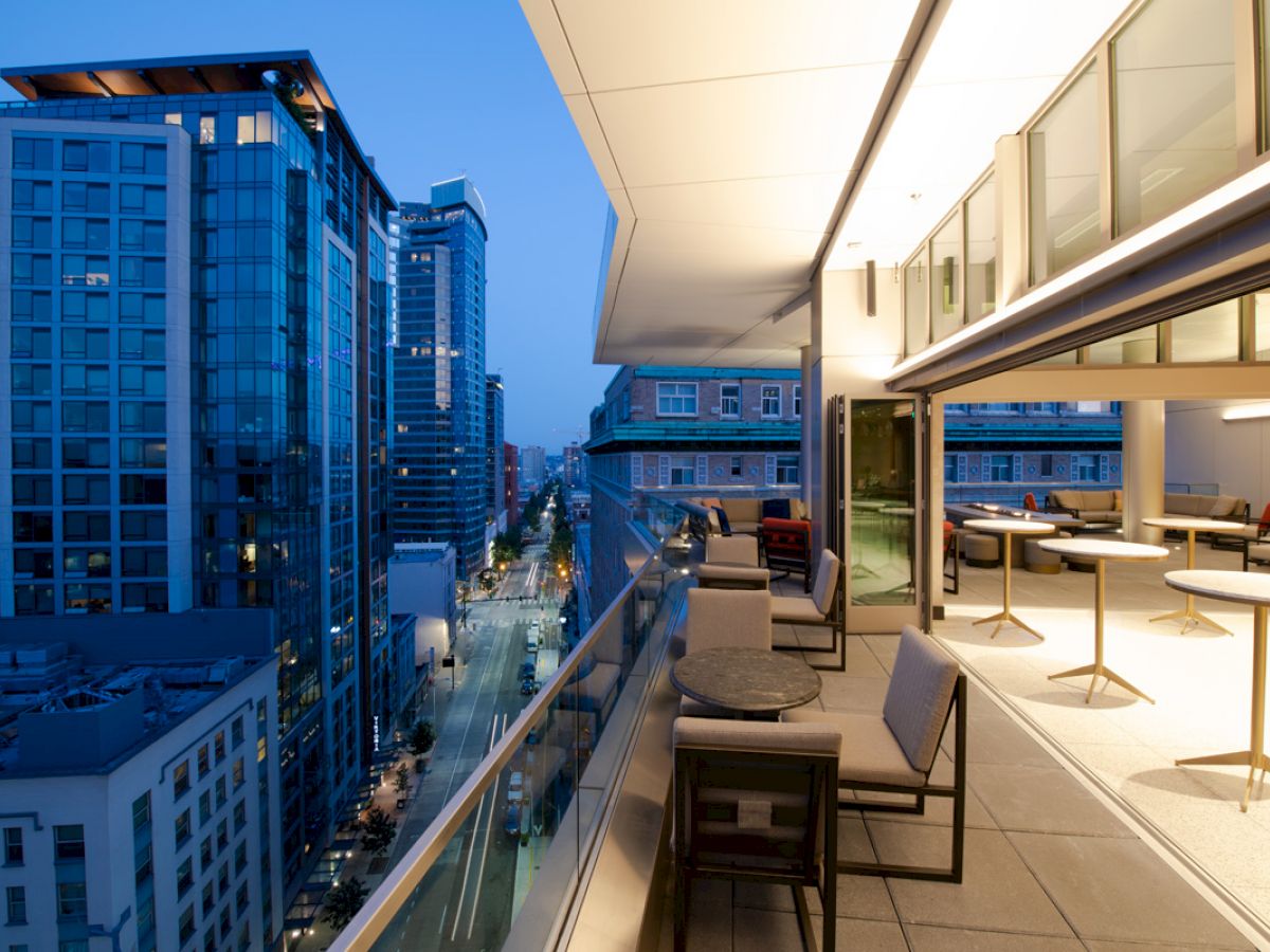 A modern balcony overlooking a city street with high-rise buildings, featuring outdoor seating and tables under soft evening light.