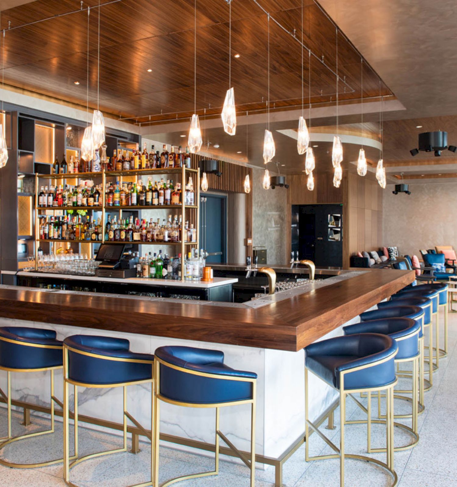 The image shows a modern bar with a well-stocked shelf, blue bar stools with gold accents, elegant lighting, and seating areas in the background.