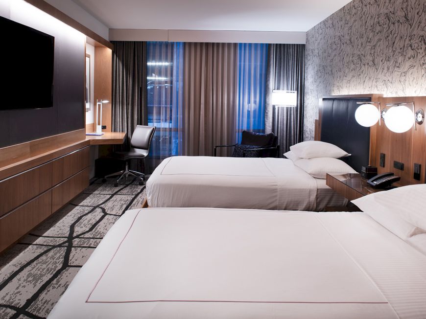 A hotel room with two beds, a wall-mounted TV, a work desk, chair, and modern lighting. The room has floor-to-ceiling windows.