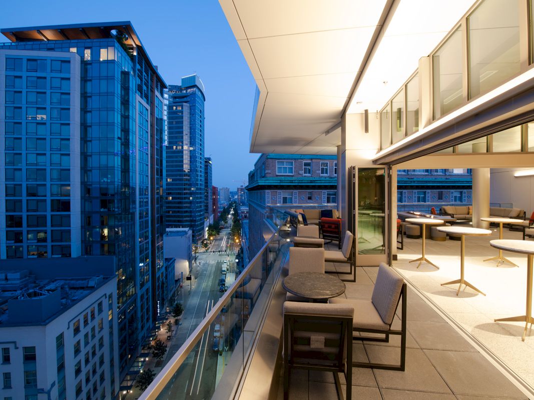 This image shows a modern balcony with tables and chairs overlooking a cityscape with tall buildings at dusk.