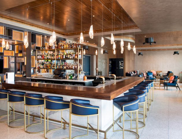 This image shows a modern bar with a central counter, various bottles on shelves, elegant lighting, and seating arrangements for patrons.