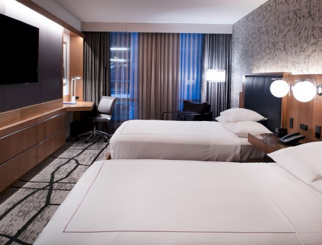 A modern hotel room with two neatly made beds, wall-mounted TV, desk, chair, and large window with curtains.