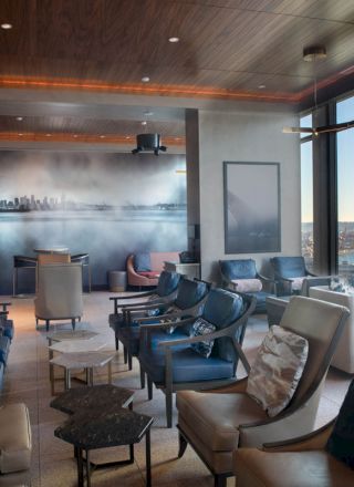 The image shows a modern lounge with several seating areas, large windows showcasing a water view, and a wall mural of a city's skyline.