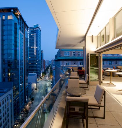 A modern cityscape view from a rooftop patio with tables and chairs, featuring tall buildings and a street lined with lights at dusk.