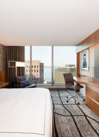 A modern hotel room with a large bed, a desk with a chair, a lounge chair, a flat-screen TV, and a large window overlooking buildings.