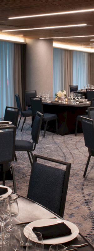 The image shows a well-lit banquet hall with round tables and chairs, elegantly set up with dishes, glassware, and floral centerpieces.