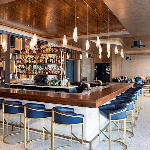 This image shows a modern bar with blue-cushioned stools, a well-stocked bar area, hanging lights, and lounge seating in the background.