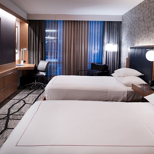 A modern hotel room with two beds, a wall-mounted TV, a work desk with a chair, and stylish decor including patterned wallpaper and curtains.