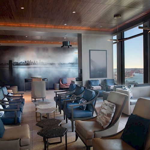 A modern lounge area with numerous comfortable chairs, large windows showcasing a waterfront view, and a mural of a city skyline on the wall.