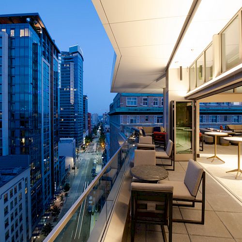 A rooftop terrace with tables, chairs, and a city skyline view at dusk. Modern buildings and illuminated streets are visible in the background.