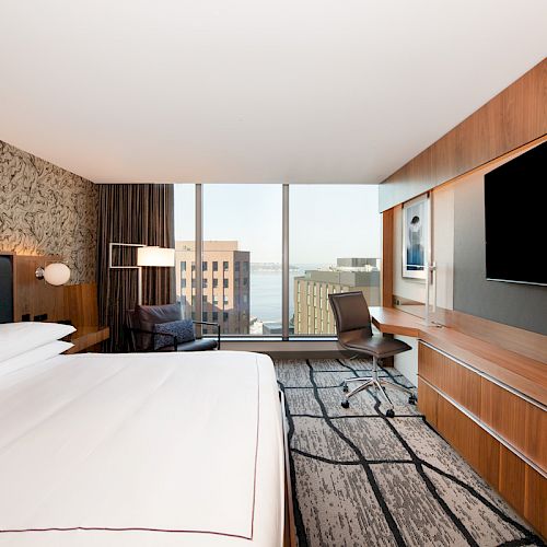 A modern hotel room features a large bed, desk with chair, TV, and a window with a city view. Decor is in neutral tones.