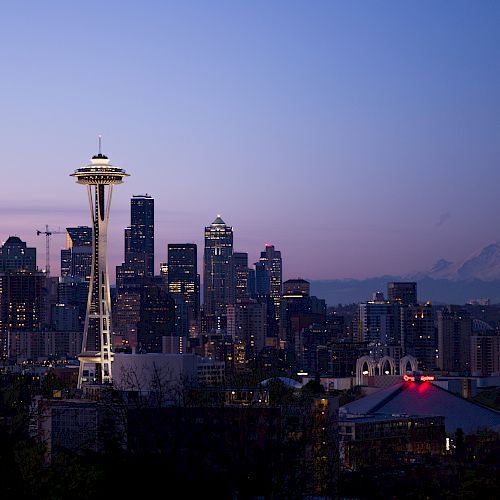 The image shows the Seattle skyline at dusk, featuring the Space Needle and Mount Rainier in the background with the city's buildings lit up.