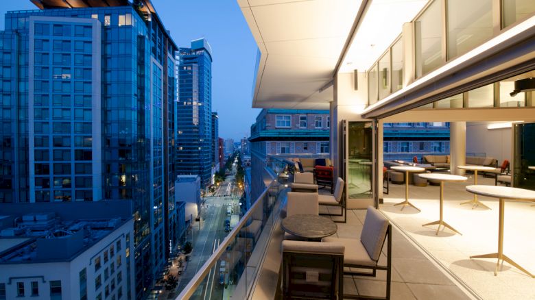 A modern cityscape view from a balcony at dusk, displaying tall buildings, a street below, and outdoor seating on the terrace.