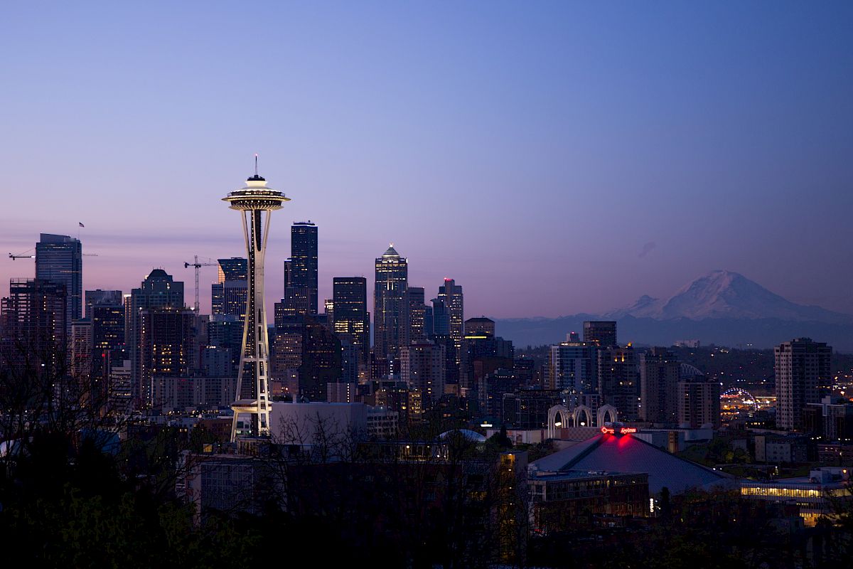 The image shows the Seattle skyline at dusk, with the Space Needle prominently displayed and Mount Rainier visible in the background.