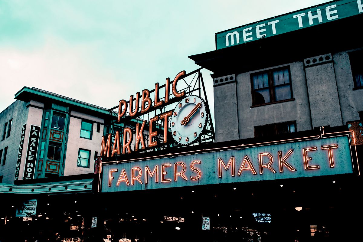 The image shows the signage of a public market with a large clock and a neon sign reading "Farmers Market." The scene appears urban and bustling.