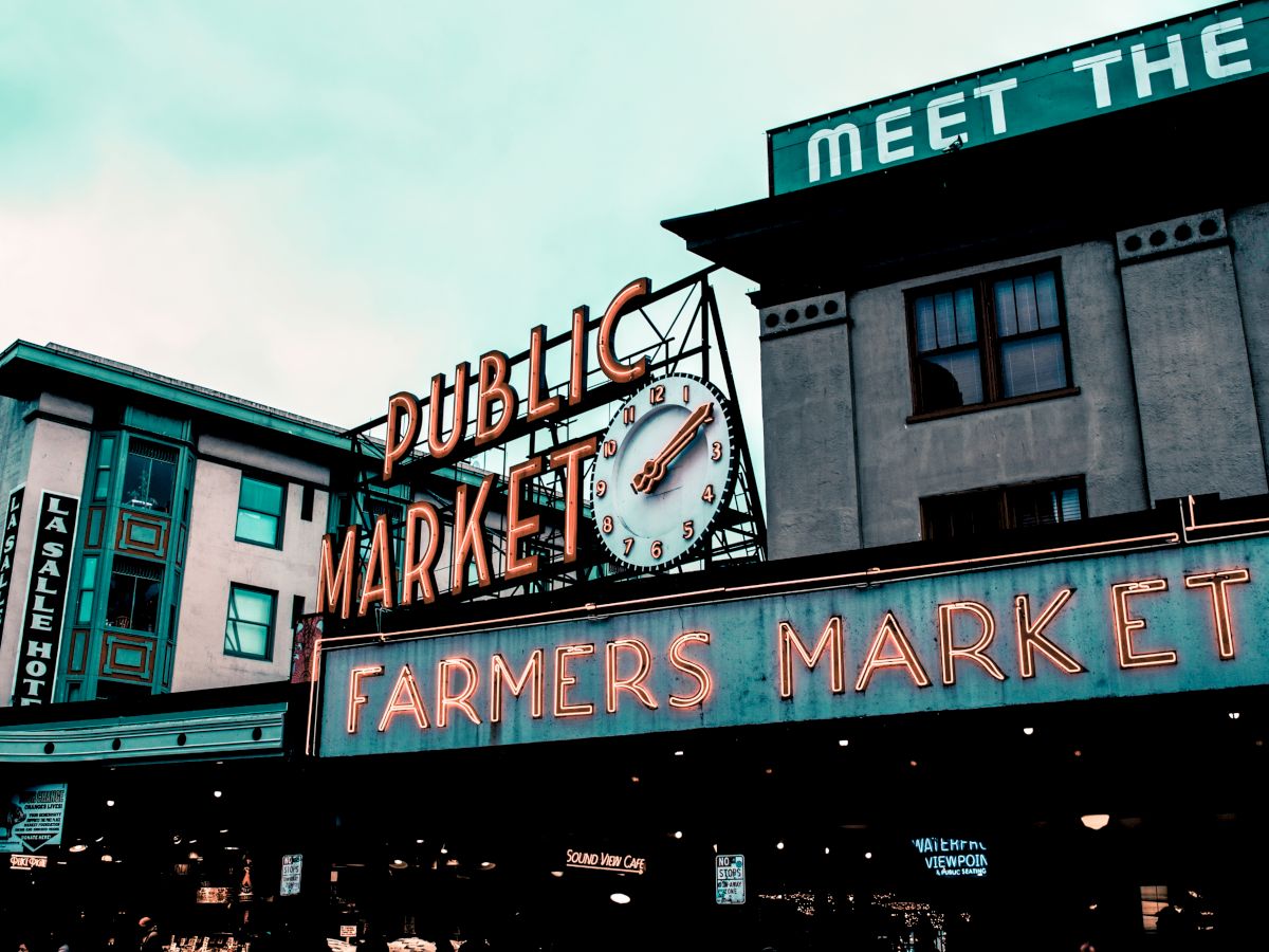 The image shows the signage of a public market with a large clock and a neon sign reading "Farmers Market." The scene appears urban and bustling.