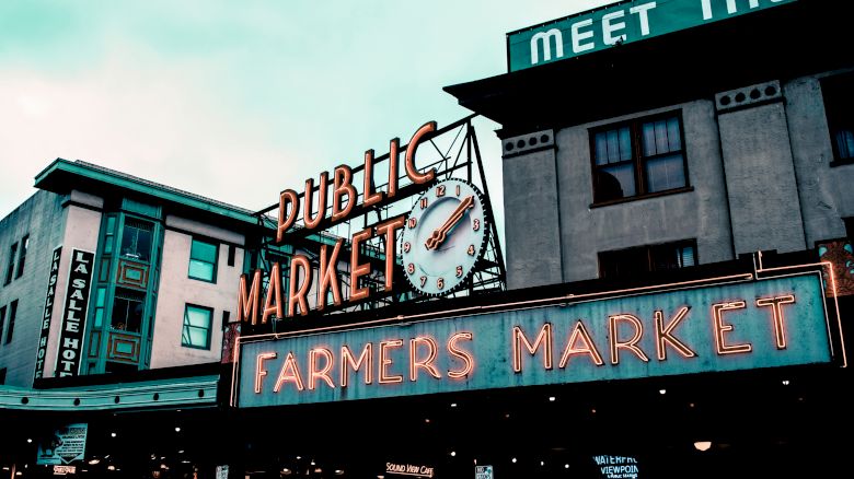 The image shows the signage of a public market with a large clock and a neon sign reading "Farmers Market." The scene appears urban and bustling.