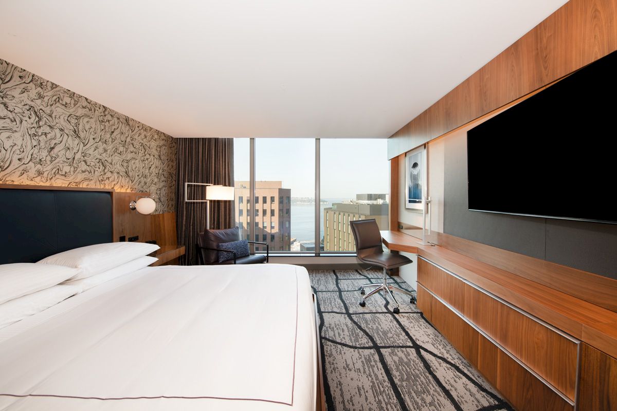 A modern hotel room with a large bed, a desk, a chair, a flat-screen TV, and a window with an urban cityscape view.