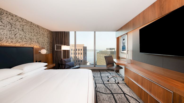 A modern hotel room with a large bed, a desk, a chair, a flat-screen TV, and a window with an urban cityscape view.
